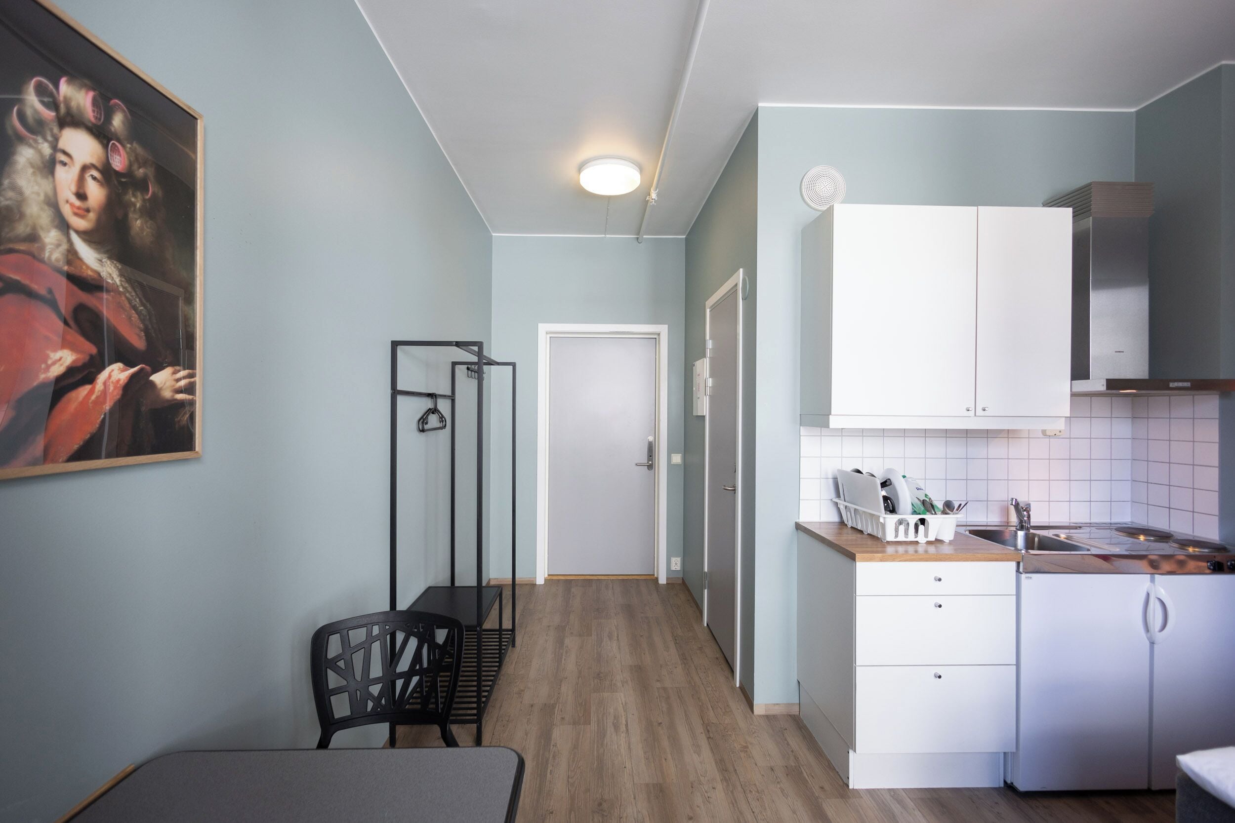A narrow hallway leading to a kitchen with white cabinets, a dining area with a table and chairs, and a painting on the wall.