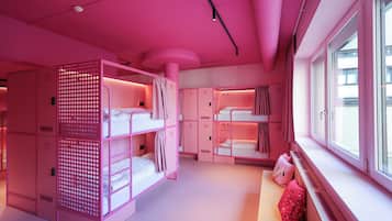 A pink-themed hotel room with bunk beds, a seating area by the window, and a large closet with numbered compartments.