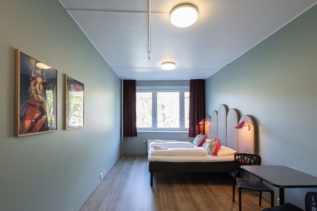 Una habitación con una cama, una mesa y una silla. Hay dos ventanas con cortinas, un radiador y luces fijadas a la pared.