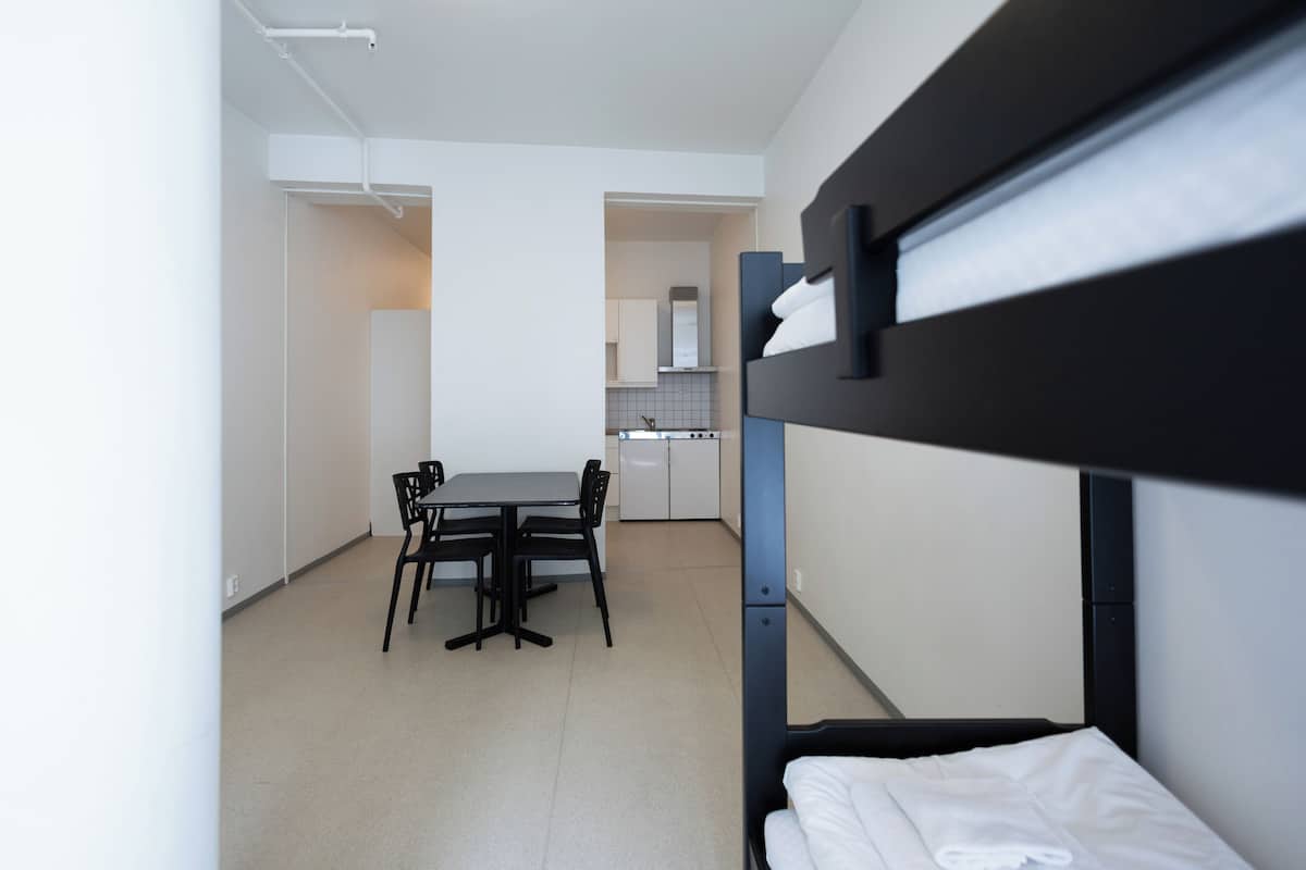 Una cama litera con un almohadón blanco, una mesa de comedor con sillas y una kitchenette al fondo.
