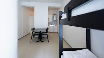 Una cama litera con un almohadón blanco, una mesa de comedor con sillas y una kitchenette al fondo.
