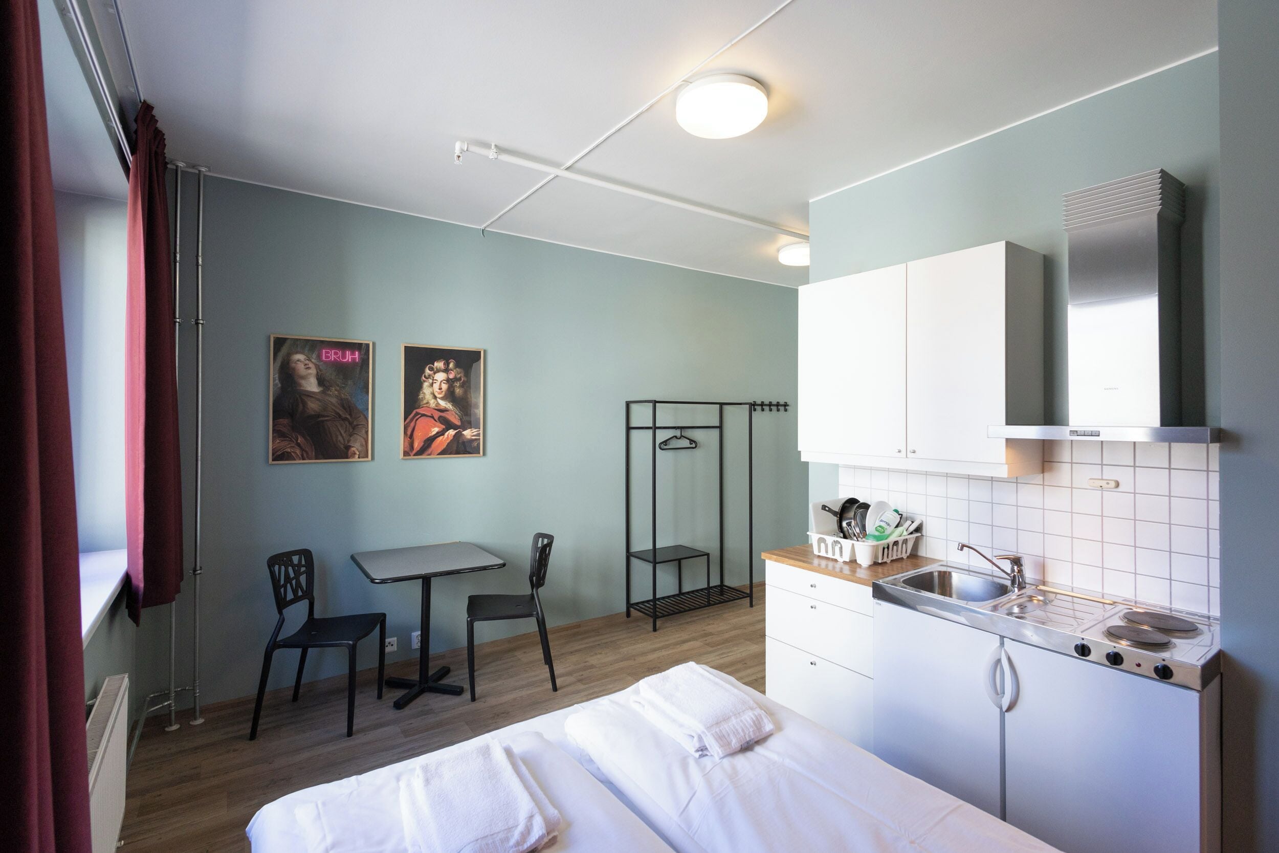 A compact hotel room with a bed, a small desk, a chair, a kitchenette, and a wardrobe.