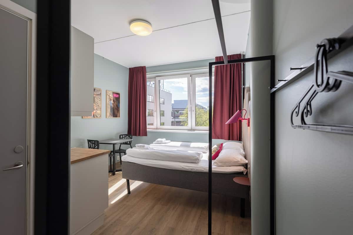 Un dormitorio con una cama, un escritorio y una silla. Hay una ventana con cortinas, un perchero en la pared y una puerta.