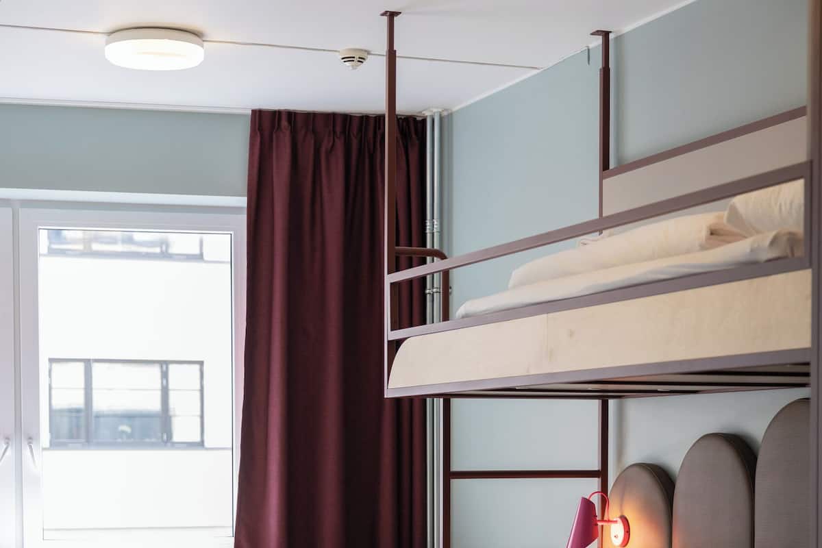Una cama litera con mesita de noche, lámpara y ventana con cortinas.