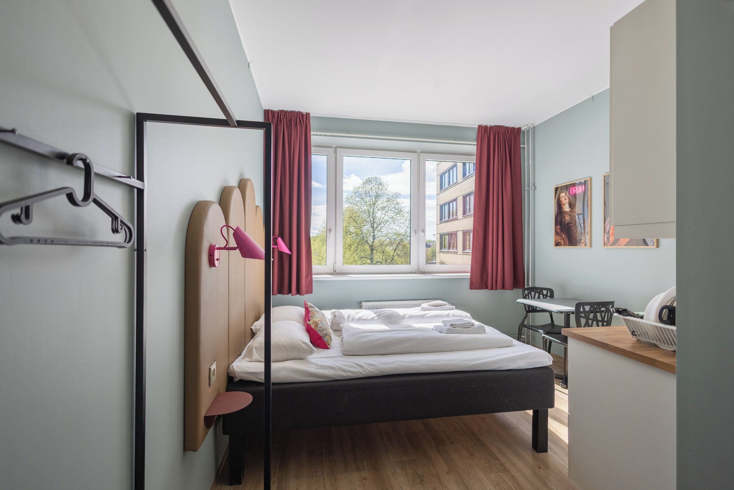 Een moderne hotelkamer met een bed, een bureau, een stoel, een raam met gordijnen en uitzicht op gebouwen.