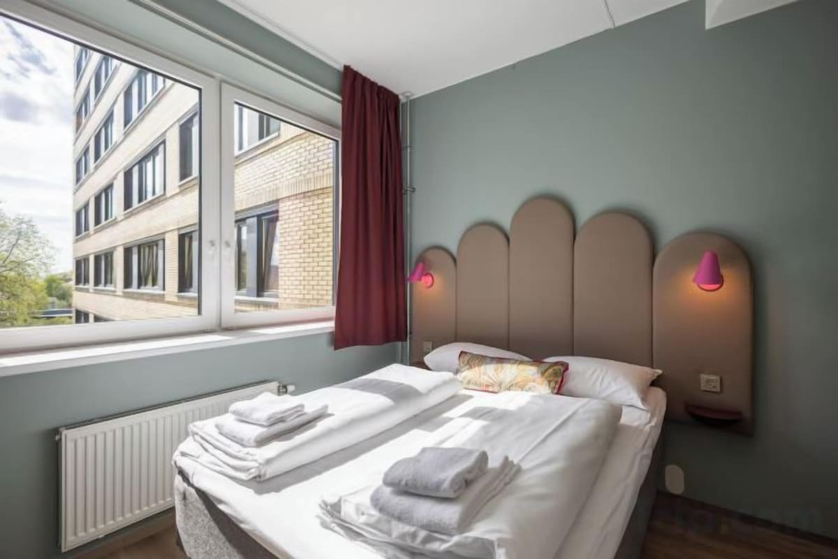 Una habitación de hotel con una cama, un ventanal, un radiador y una lámpara fijada a la pared.