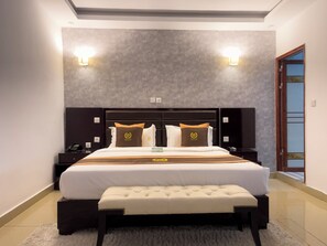 A modern hotel room with a large bed, two bedside tables, a bench, and a wall-mounted light fixture. - Hôtel Sana (Abidjan)
