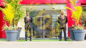 Two men in suits standing in front of the SANA HOTEL entrance, with potted plants and a red building facade. - Hôtel Sana (Abidjan)