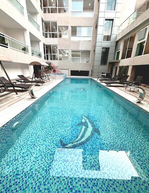 An indoor swimming pool with tiled mosaic patterns, surrounded by lounge chairs and a modern building with balconies. - Hôtel Sana (Abidjan)