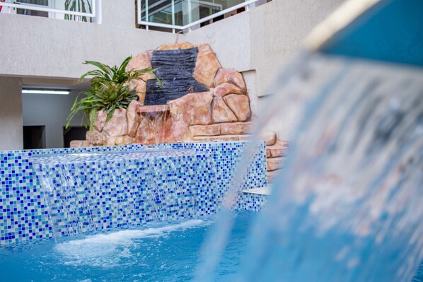 A swimming pool with a waterfall feature, surrounded by a tiled wall and a potted plant.