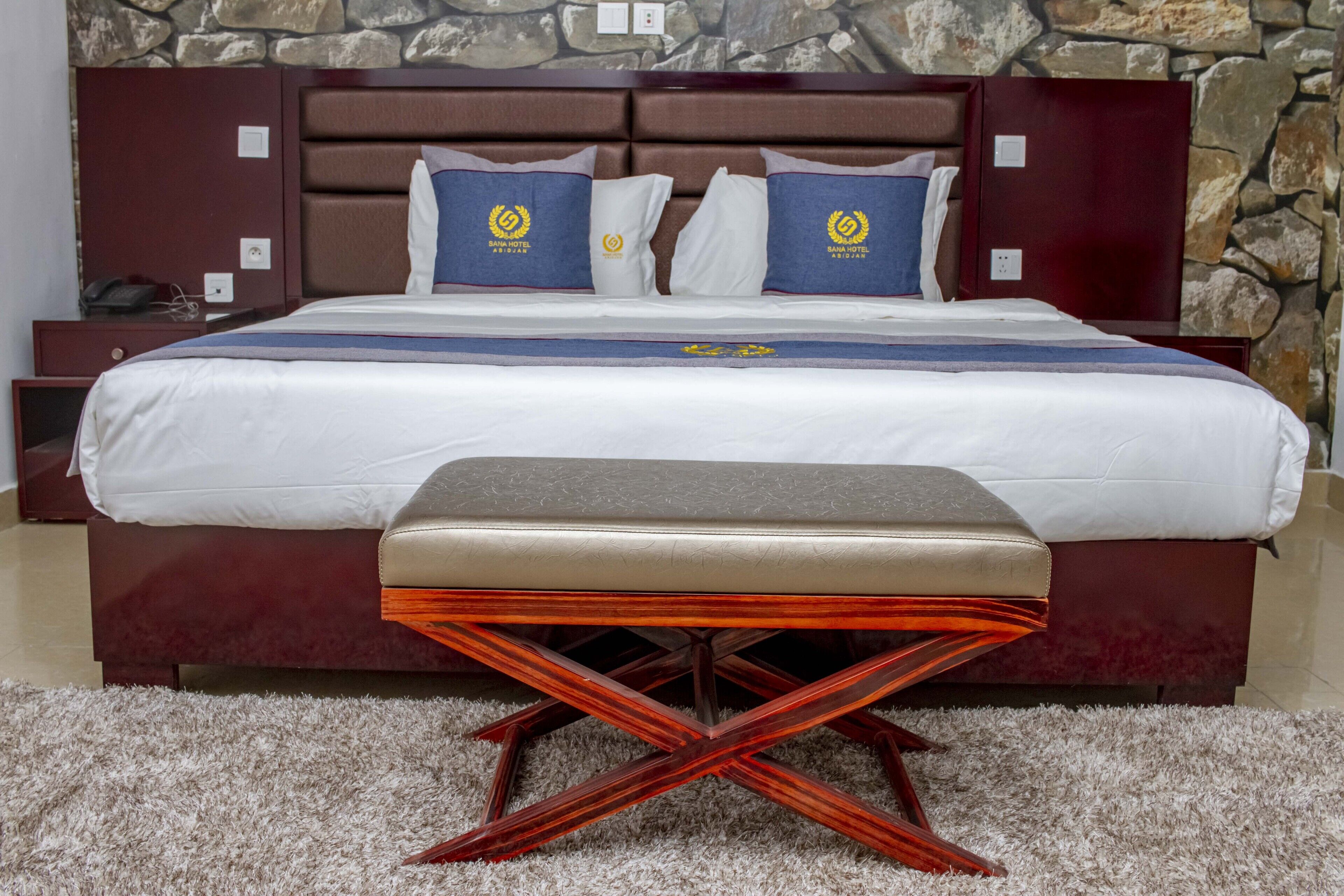 A hotel room with a bed, a bench, a nightstand, and a stone wall.