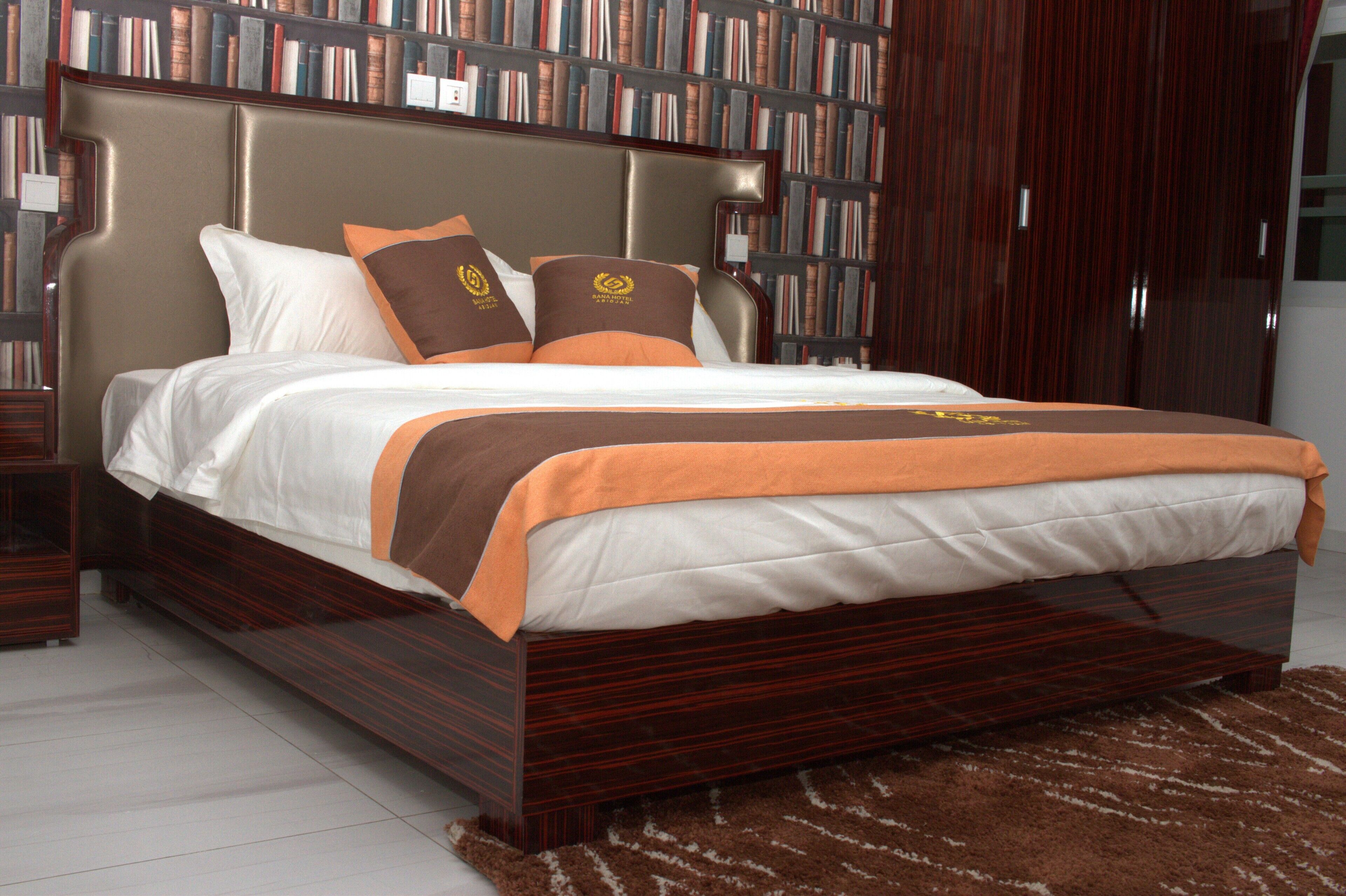 A large bed with a wooden frame, white and orange bedding, and two pillows with a crest emblem.