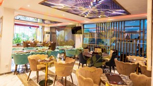 A modern restaurant with a variety of seating arrangements, including round tables and individual chairs, with a view of the outdoors through large windows. - Hôtel Sana (Abidjan)