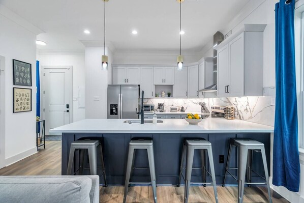 Una cocina moderna con armarios blancos, una isla grande con taburetes, electrodomésticos de acero inoxidable e iluminación colgante.
