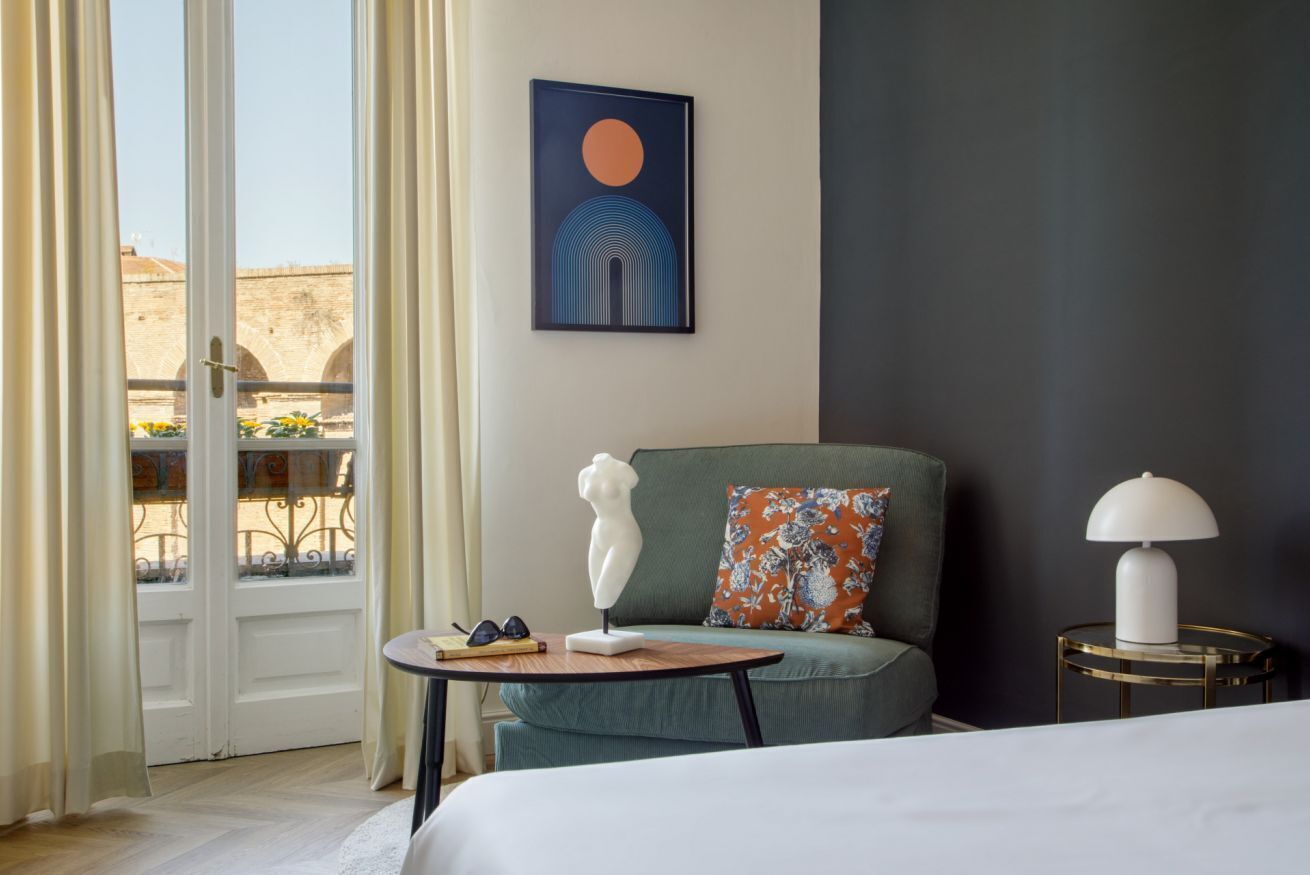 A hotel room with a bed, a sofa, a table with a lamp, a painting on the wall, and a view of the outside.