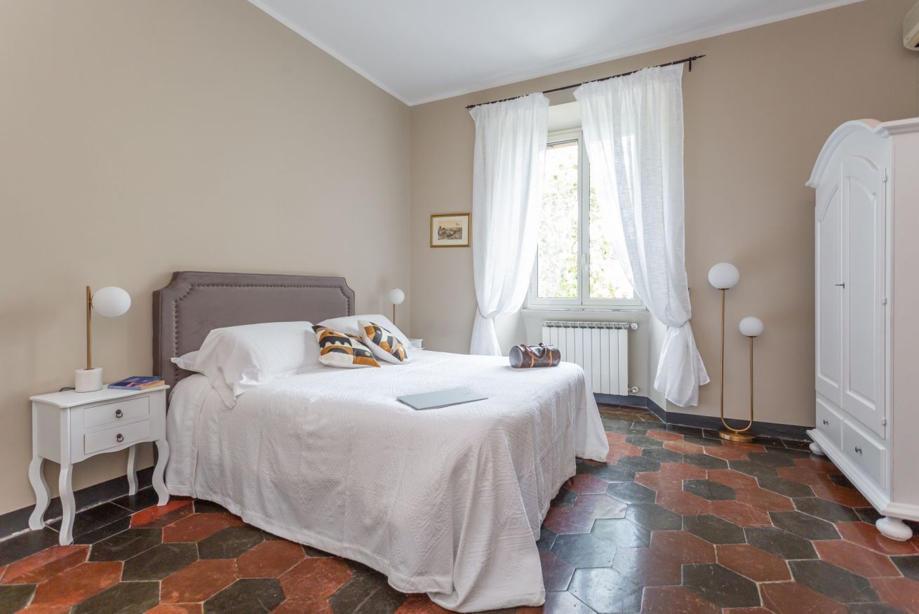 Een slaapkamer met een bed, witte gordijnen, een radiator, een kledingkast en een nachtkastje met een lamp.