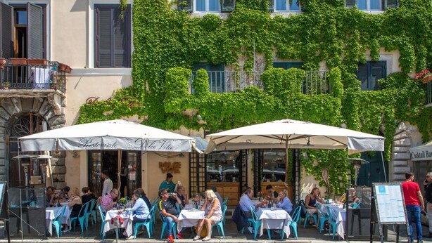 Un café en plein air, avec des tables et des chaises, entouré d’un bâtiment orné de plantes grimpantes vertes et doté de volets bleus.
