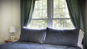 A bed with a quilted blue comforter and two pillows, a bedside table with a lamp and an alarm clock, and a window with green curtains. - The Coastal Cottage | Walk to Pemaquid Beach | Fire Pit & Family-Friendly Retreat (Bristol)