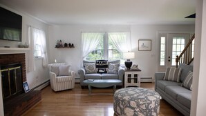 A living room with a fireplace, two sofas, a coffee table, and a patterned ottoman. - The Coastal Cottage | Walk to Pemaquid Beach | Fire Pit & Family-Friendly Retreat (Bristol)