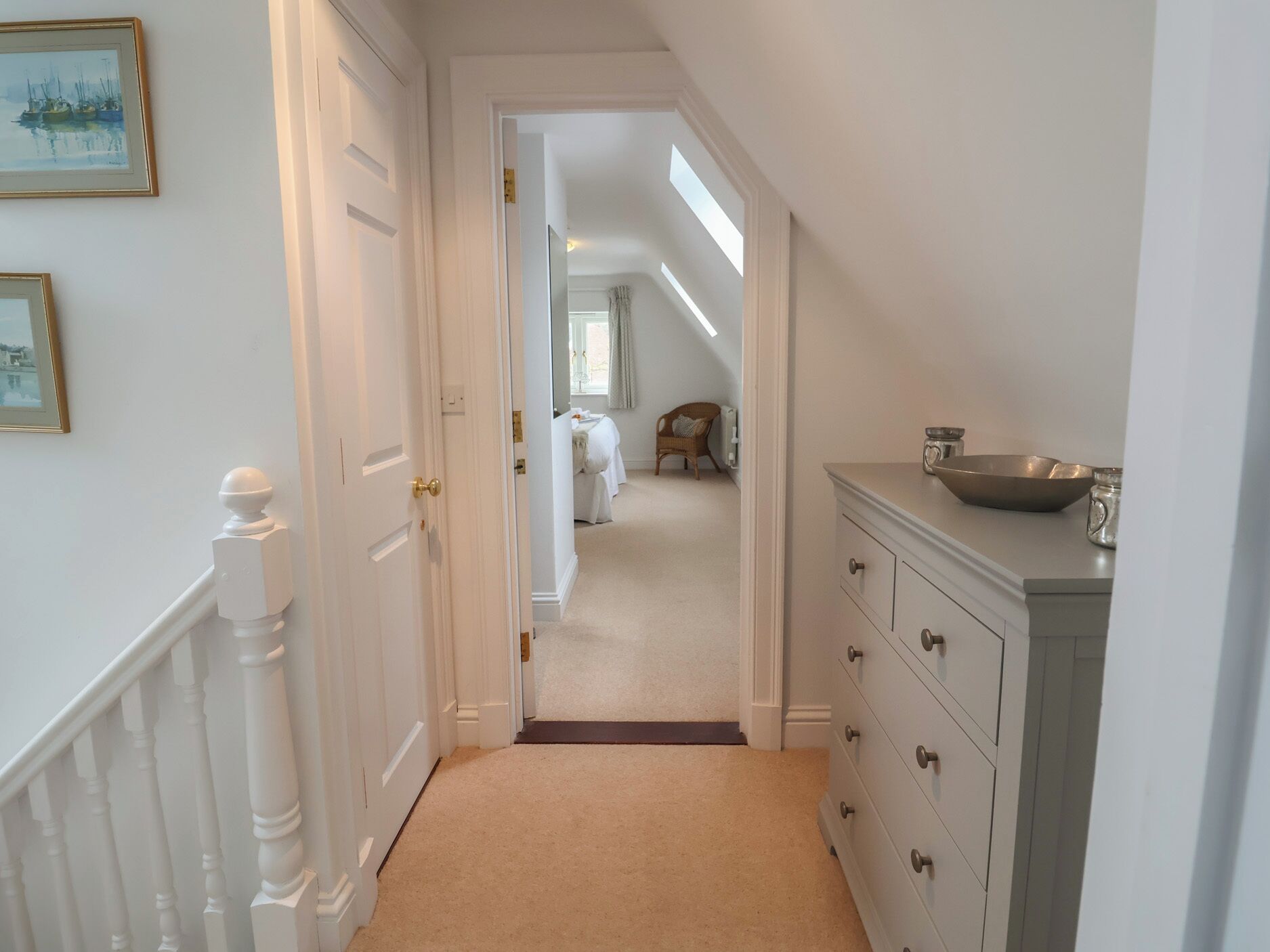 A hallway with a white door, a staircase with white balusters, a beige carpet, a white dresser with drawers, and a painting on the wall.