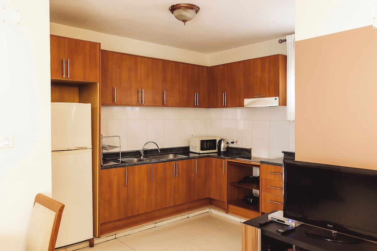 A compact kitchen with wooden cabinets, a microwave, a refrigerator, a sink, and a flat-screen TV.