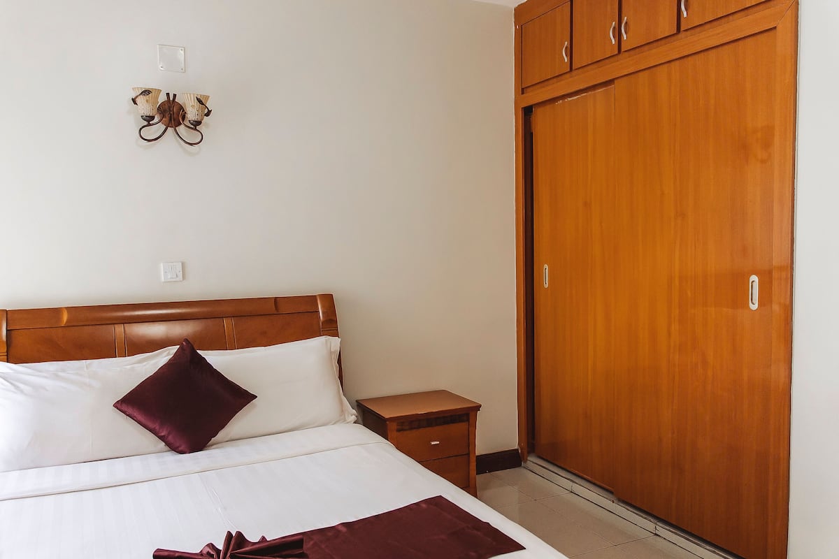 A hotel room with a wooden headboard, a single bed with white linens and a maroon pillow, a wooden nightstand, a wooden wardrobe, and a wall-mounted light fixture.