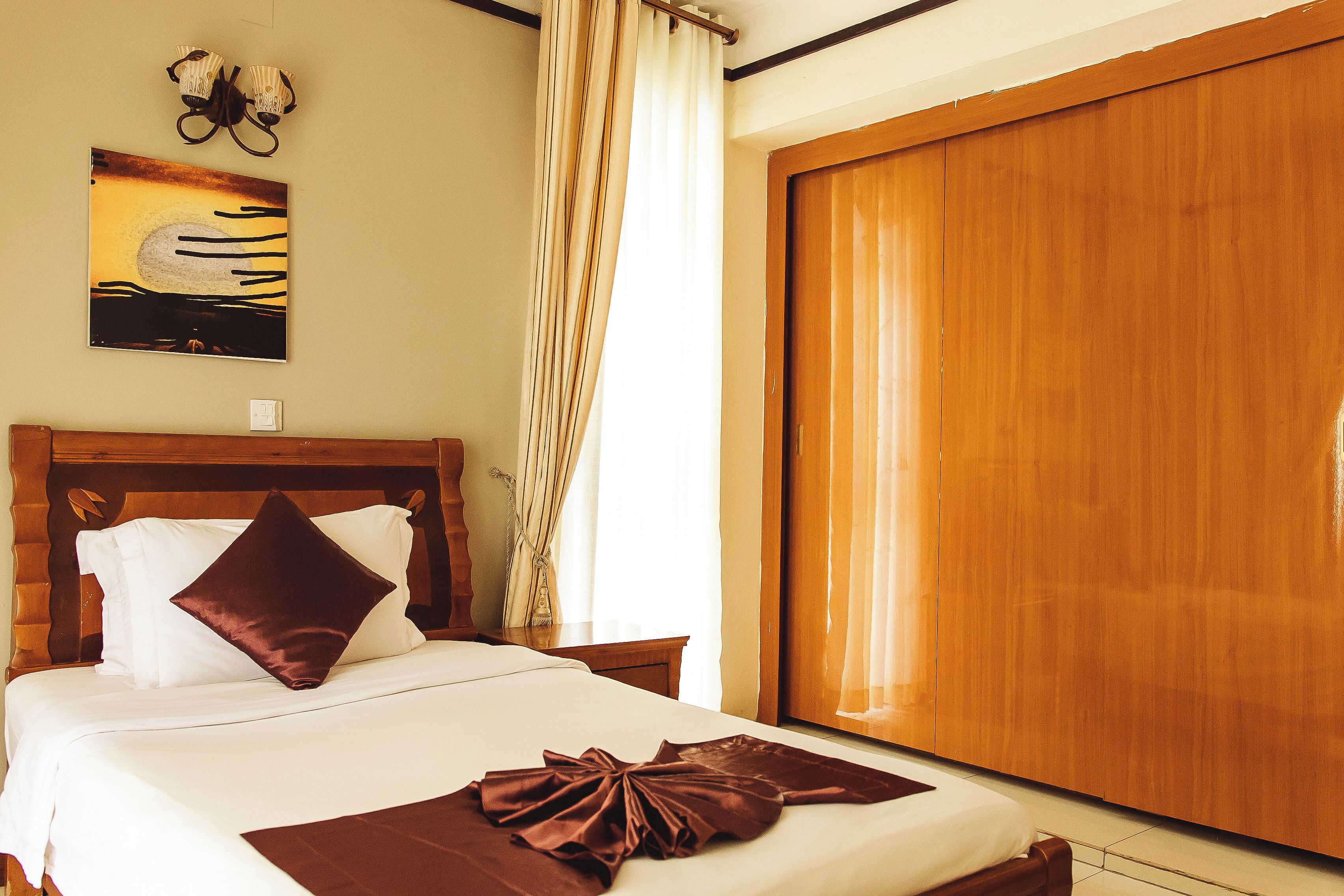 Habitación de hotel con cabecera de madera, una cama grande con ropa de cama blanca y marrón, un plaid marrón sobre la cama, un armario de madera y un cuadro en la pared.