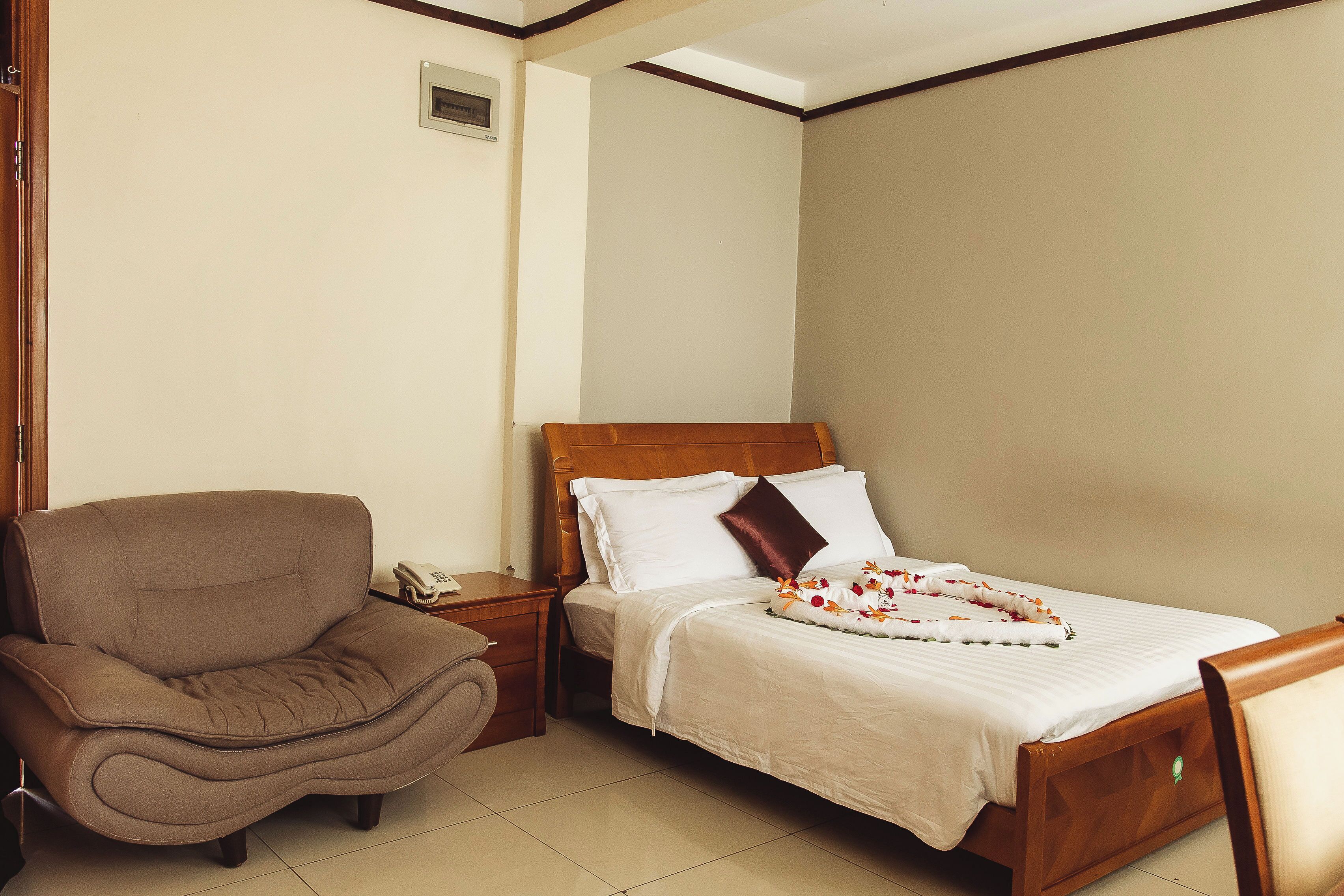 A hotel room with a bed, a wooden nightstand, a brown armchair, and a small bedside table with a telephone.