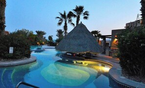 A resort pool area with a thatched-roof structure, palm trees, and a clear blue pool.