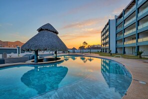 A resort pool with a thatched-roof structure, a modern building with balconies, and a sunset in the background. - Modern Condo with Ocean Views (Las Conchas)