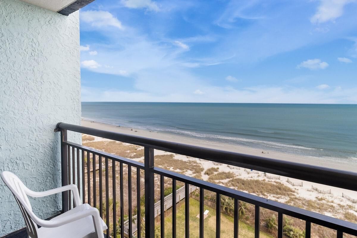 A balcony with a view of the beach, a white plastic chair, and a railing.