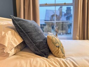 A bed with two pillows, one grey and one yellow with embroidery, in a room with a window showing a building outside. - Captivating 3-bed Cottage in Saltaire (Shipley)