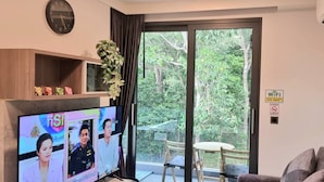 Un salon comprenant une télévision, une horloge, une plante, un tableau et une porte-fenêtre coulissante donnant sur un balcon.
