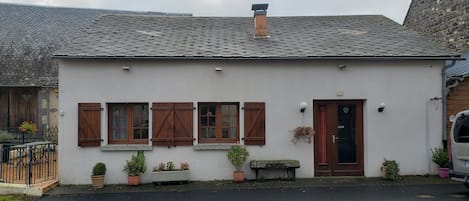 Un bâtiment blanc avec un toit en tuiles, des volets en bois et une porte ornée d’un motif en forme de cœur. On y trouve des plantes en pot et un banc à l’extérieur.