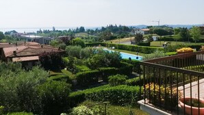 A residential area with a swimming pool surrounded by lush greenery, a clear view of the sea, and a distant cityscape. - Appartamento Giada (Padenghe sul Garda)