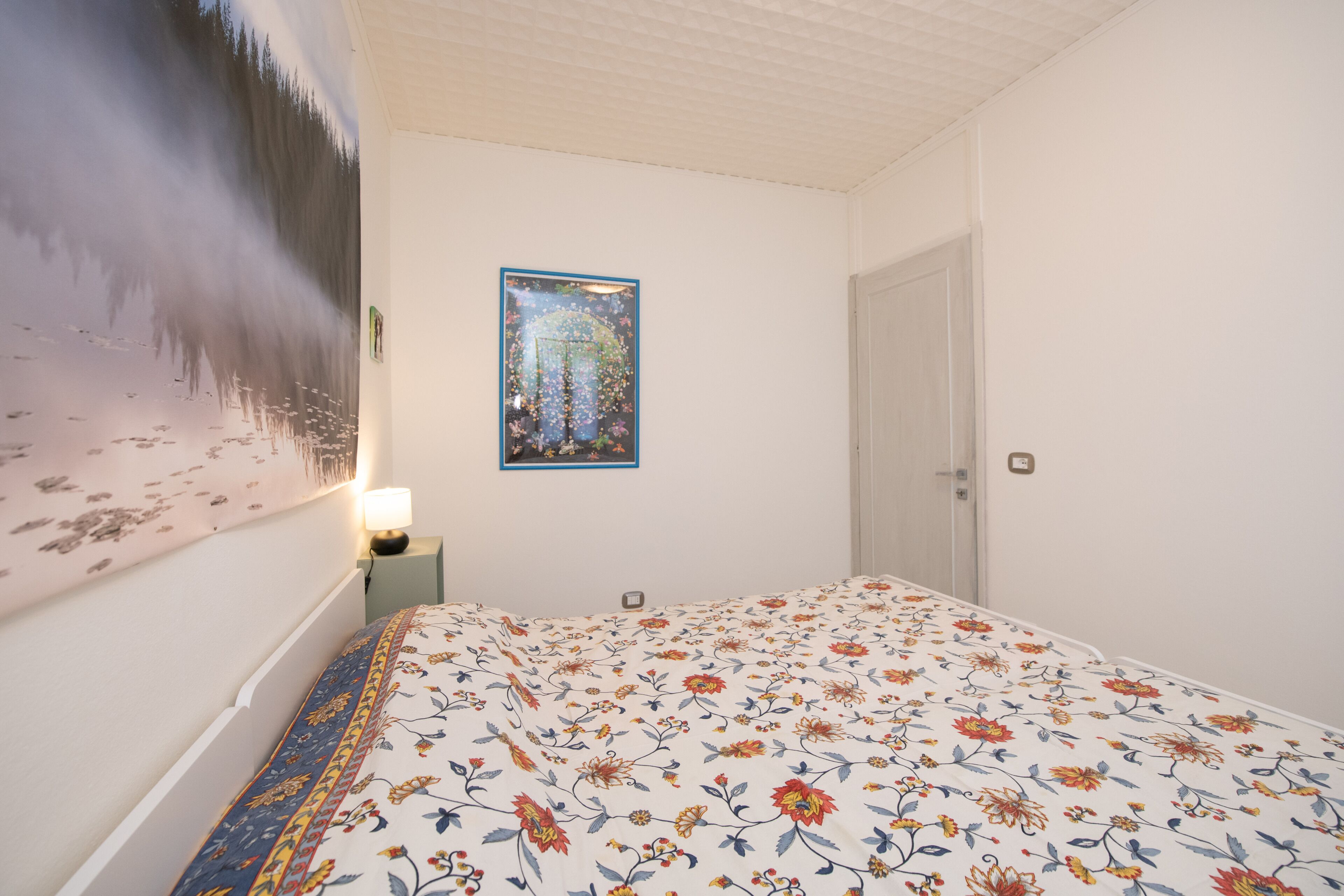 A bedroom with a floral bedspread, a wall-mounted painting, and a bedside lamp.
