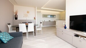 A modern living room with a grey sofa, a dining table with white chairs, a TV on a cabinet, and a kitchen area in the background. - Appartamento Giada (Padenghe sul Garda)