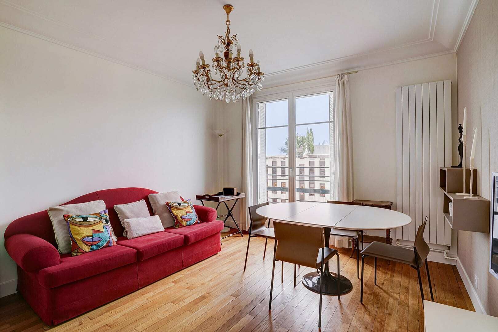 Un salon moderne avec un canapé rouge, une table ronde entourée de chaises, un lustre et une vue sur les immeubles à travers de grandes fenêtres.