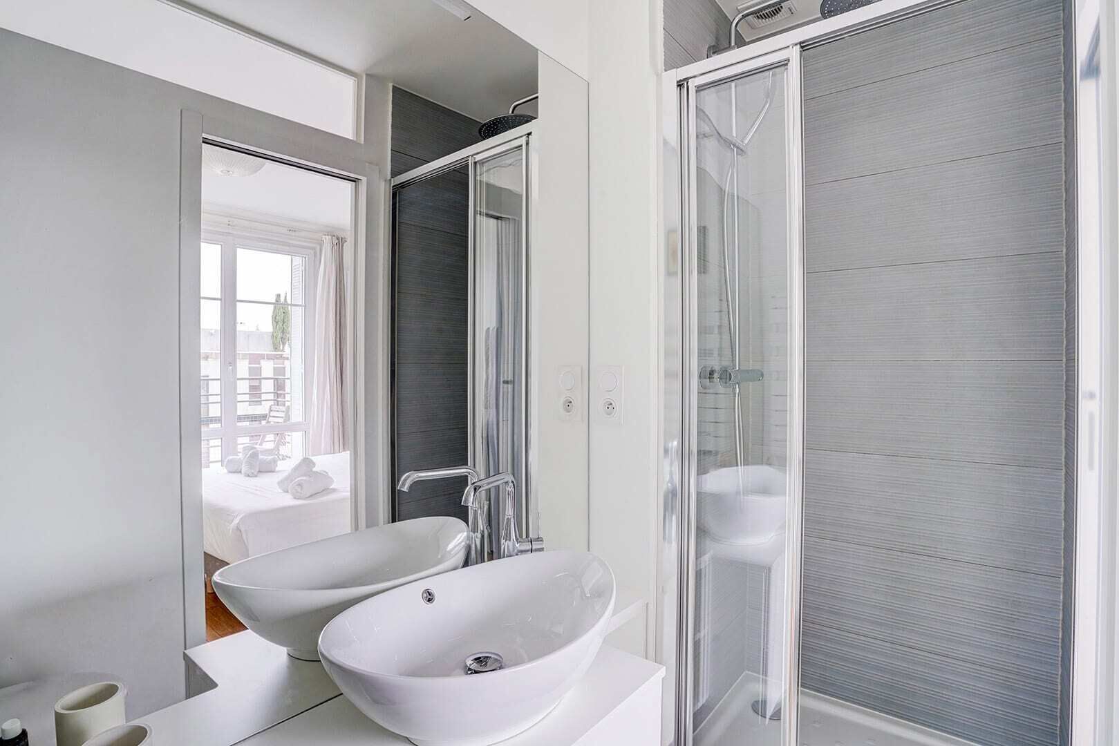 Une salle de bain moderne avec un grand miroir, deux vasques en céramique blanche, un espace douche et une vue sur le balcon extérieur.