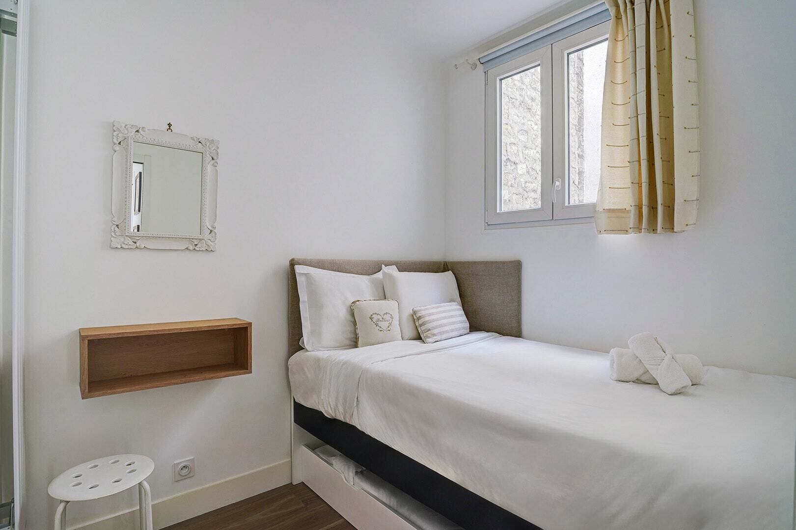 A bedroom with a bed, a bedside table, a mirror, and a window with curtains.