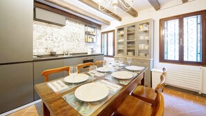 A dining area with a long wooden table set for six, flanked by chairs. A kitchen with modern appliances and a patterned backsplash is visible in the background.