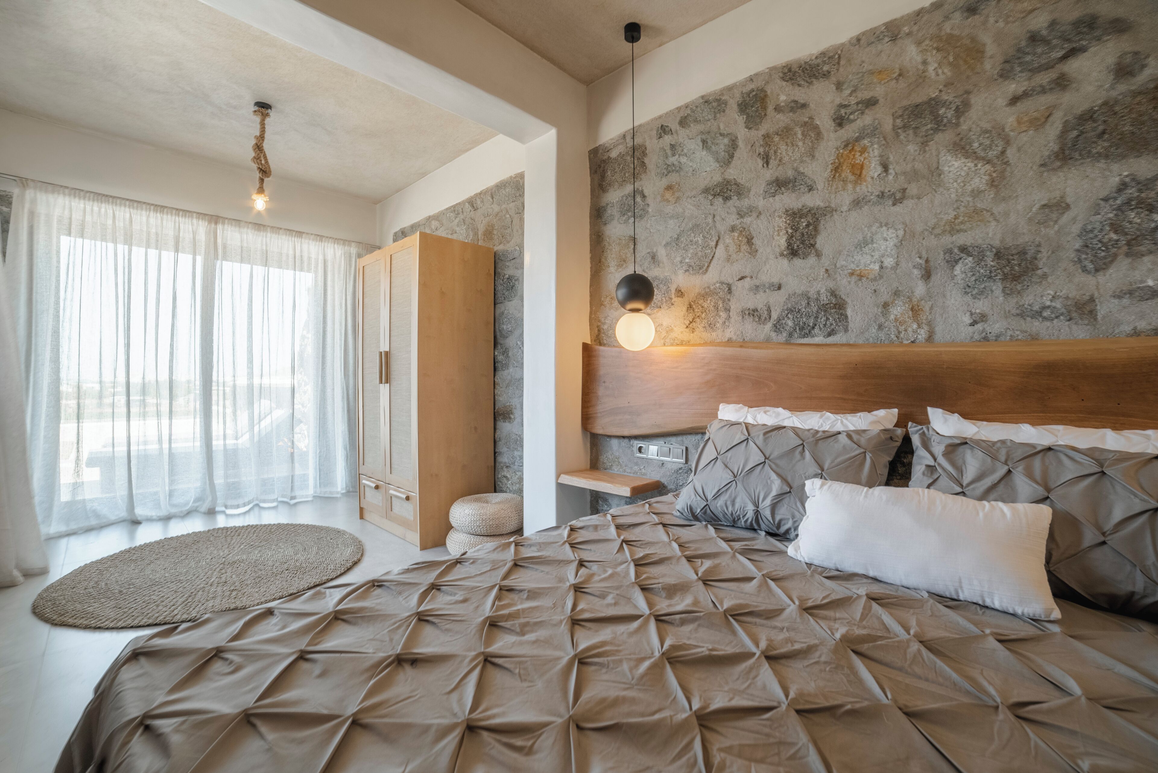 A bedroom with a large bed, a stone wall, a wooden headboard, a bedside table, and a hanging light fixture.