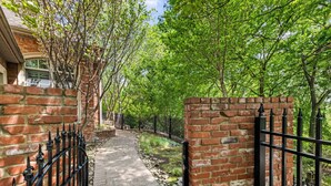A brick pathway with a metal fence, surrounded by lush green trees.
