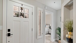Un couloir avec une porte blanche, une fenêtre avec des stores et un lit à l’arrière-plan.