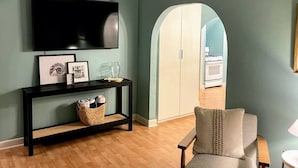 A living room with a TV, framed pictures, a console table, a chair, and an arched doorway leading to a kitchen.