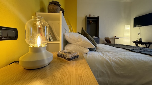 Une chambre d’hôtel avec une table de chevet en bois, une lampe, un lit avec une literie blanche, une bibliothèque et une armoire.