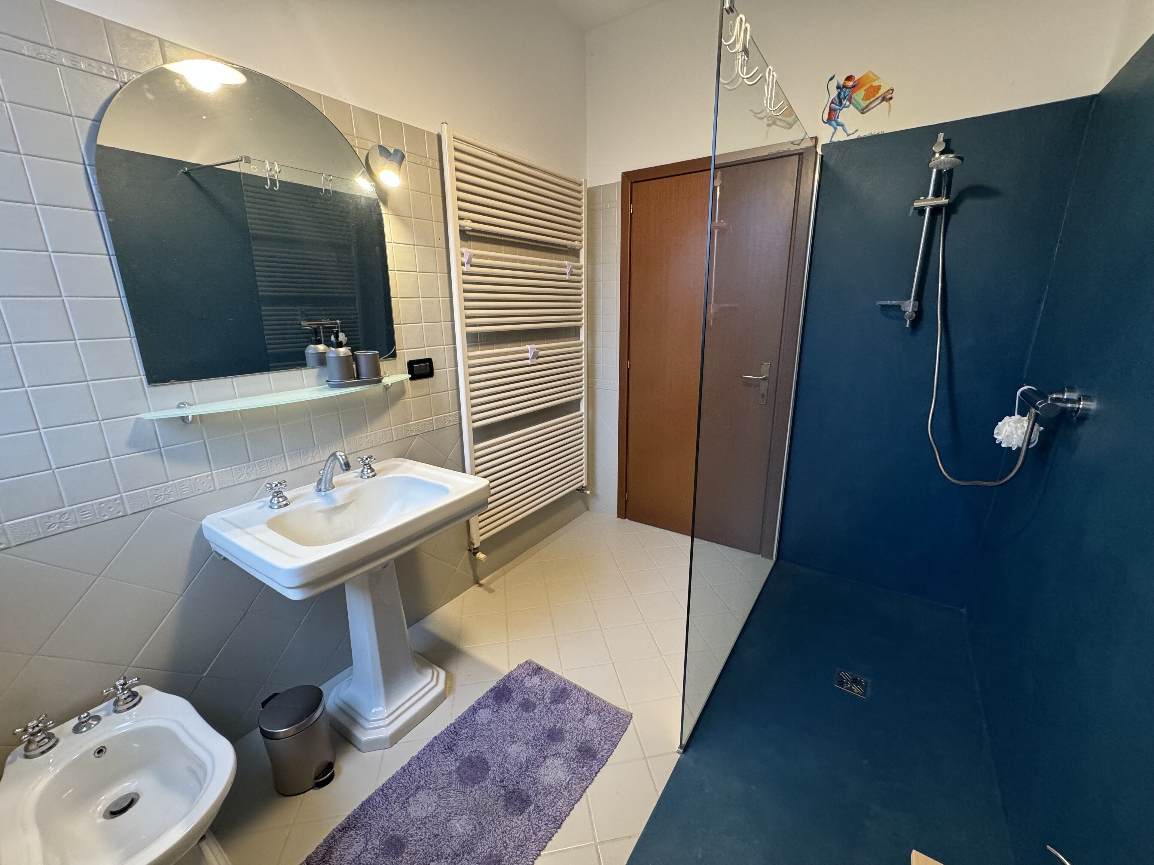 Et badeværelse med et håndvaskarrangement, et bidét, et toilet, et håndklædestativ, en bruser og et spejl.