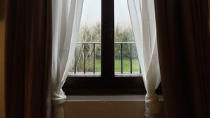 Une fenêtre ornée de rideaux blancs et légers, une croix noire au-dessus, et une vue sur un jardin verdoyant.
