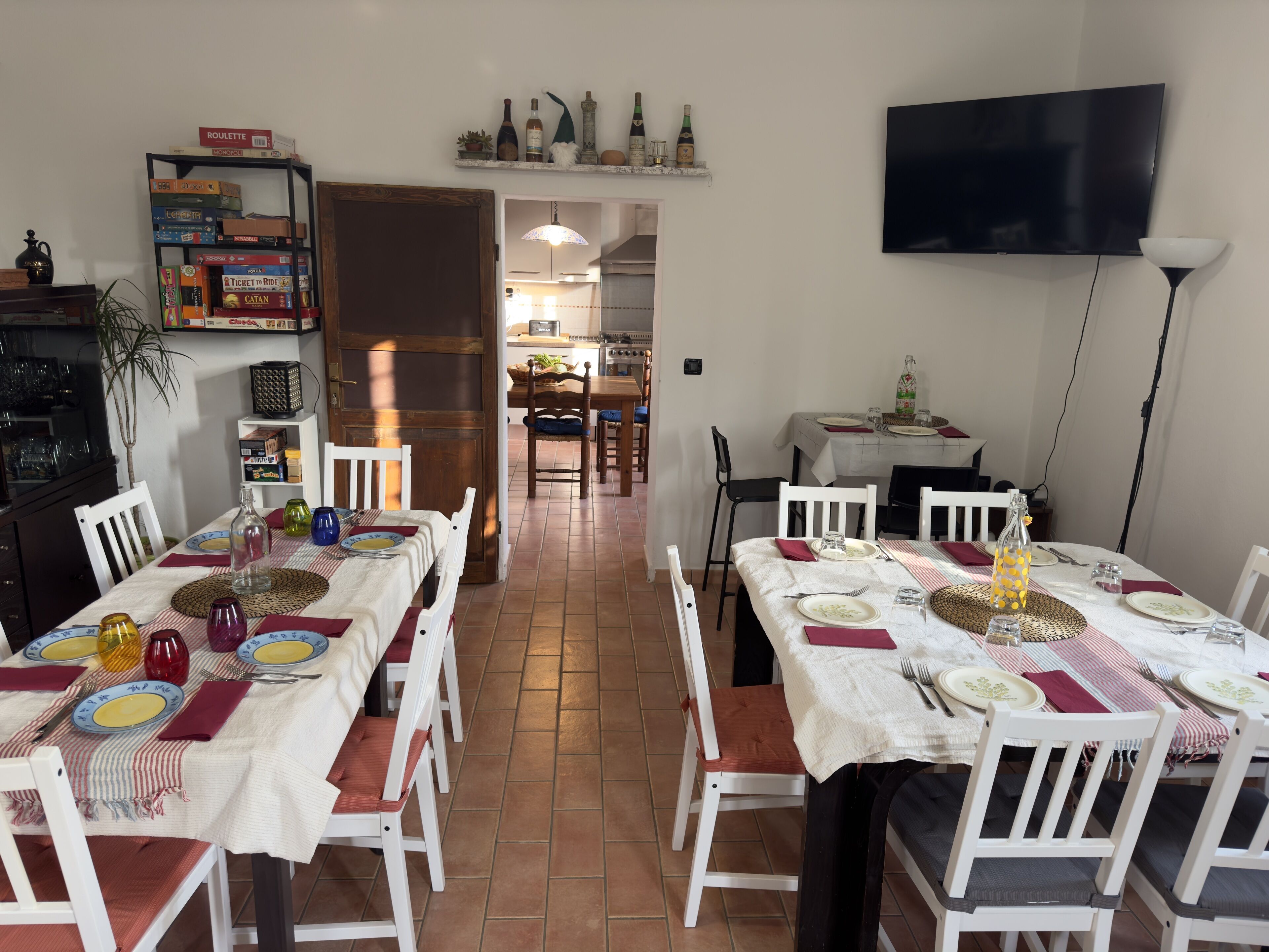 Zona pranzo con tavoli e sedie bianche, cucina sullo sfondo e televisione appesa al muro.
