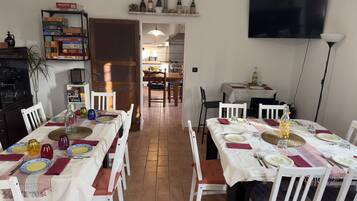 Zona pranzo con tavoli e sedie bianche, cucina sullo sfondo e televisione appesa al muro.